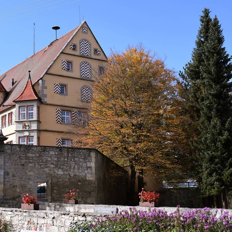 Ein historisches Gebäude mit verziertem Fachwerk und Satteldach steht hinter einer alten Steinmauer, umgeben von Bäumen und Blumenbeeten.