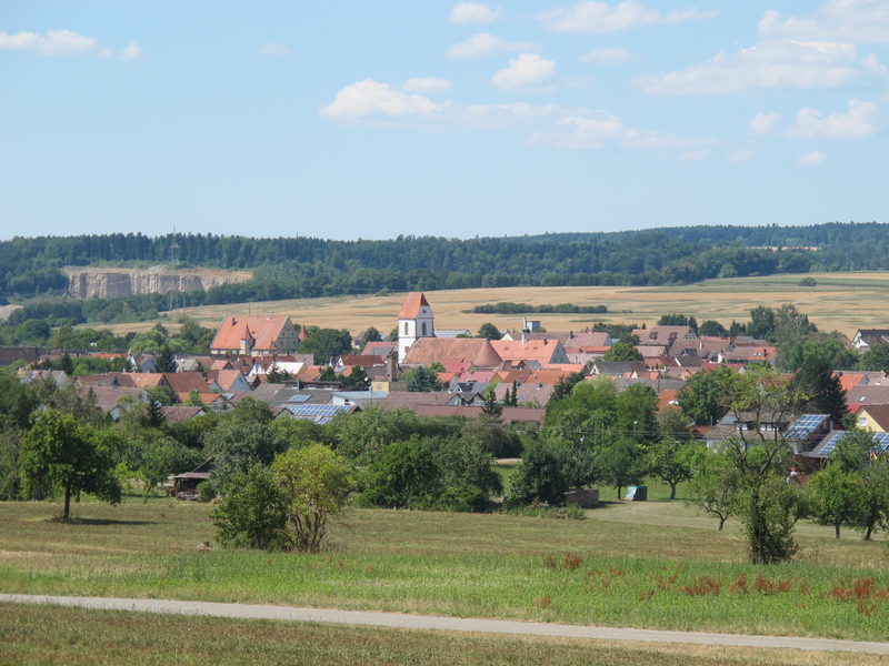 Ein Dorf mit rot-gedeckten Häusern, einer Kirche mit Turm und umliegenden Feldern und Wäldern unter einem blauen Himmel mit einigen Wolken.