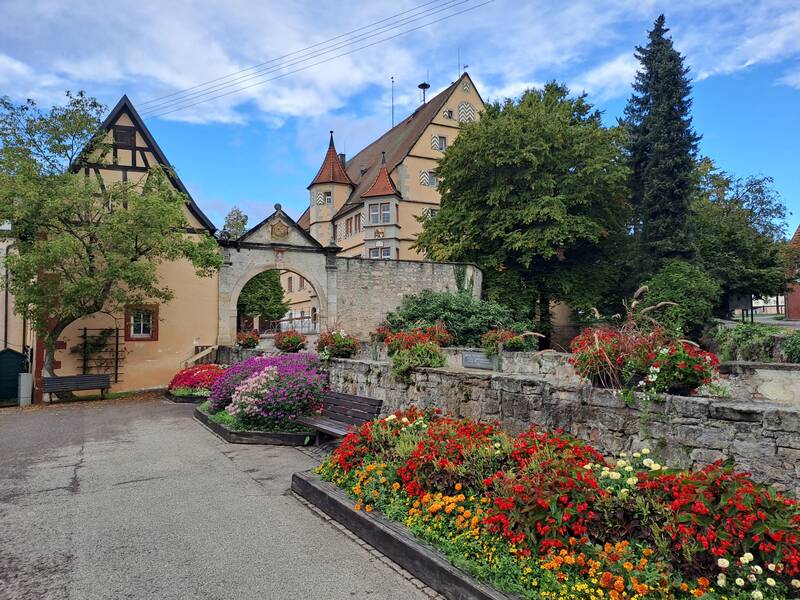 Ein malerischer kleiner Platz mit blühenden Blumenbeeten, einer historischen Steinmauer mit Torbogen und Fachwerkhäusern im Hintergrund.