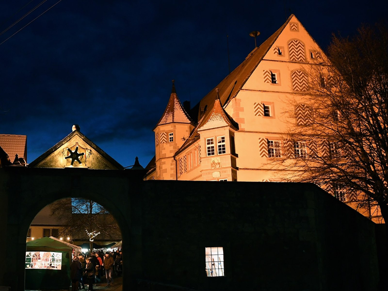 Das Bild zeigt einen beleuchteten historischen Gebäudekomplex in der Abenddämmerung, im Vordergrund befindet sich ein Torbogen mit einem kleinen hell erleuchteten Weihnachtsmarkt.