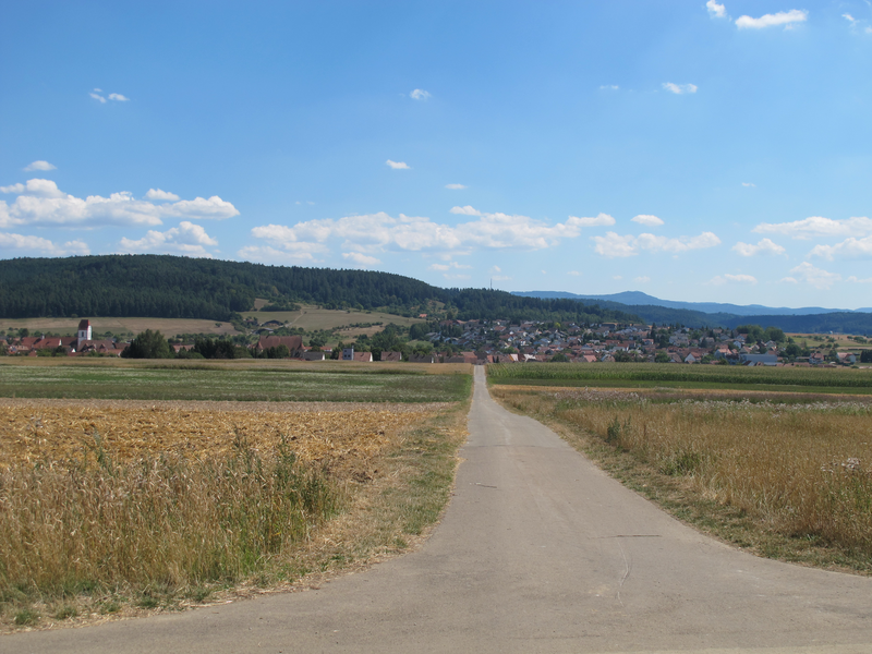 Ein schmaler Weg führt durch eine ländliche Landschaft aus Feldern und Wiesen zu einem kleinen Dorf mit Kirche vor bewaldeten Hügeln.