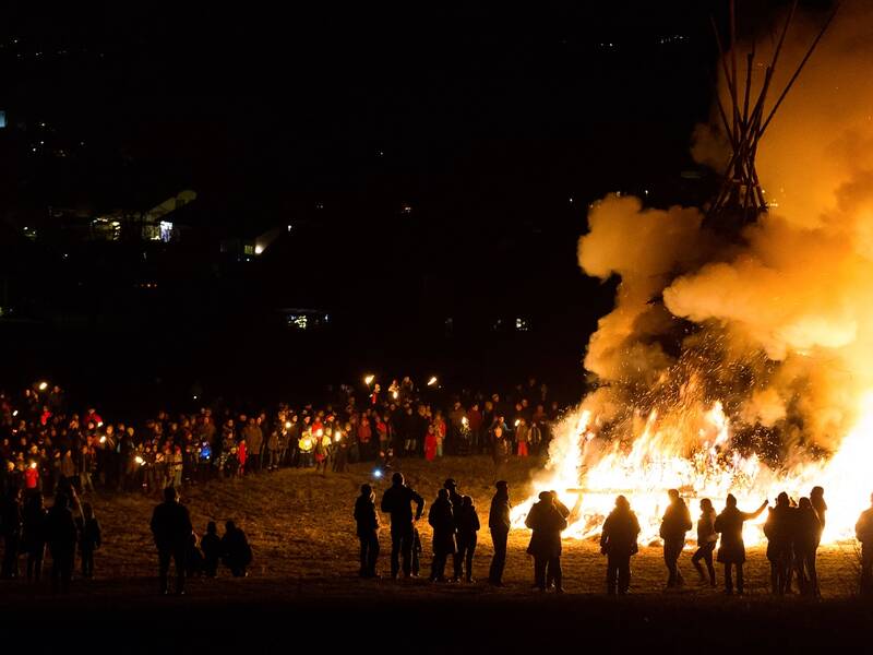 Eine große Menschenmenge versammelt sich in der Nacht um ein großes, hell loderndes Feuer.