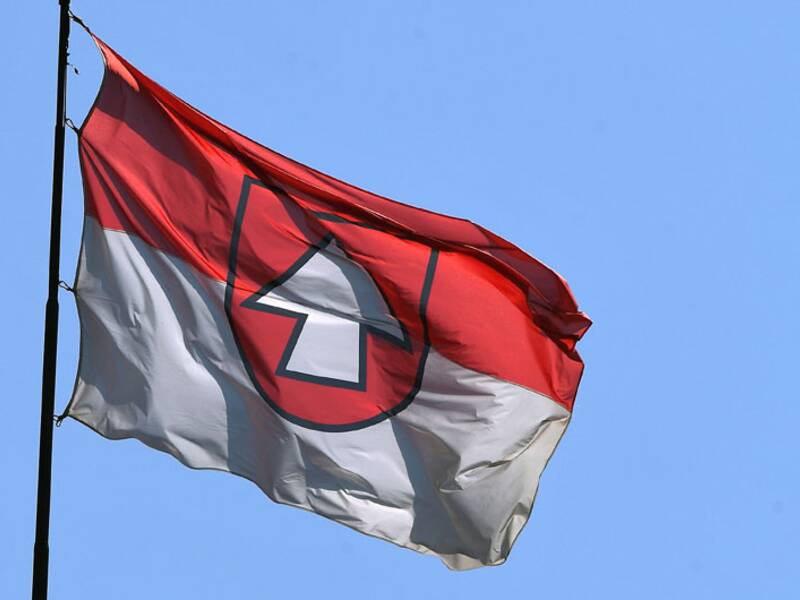 Eine Flagge mit einem roten und weißen Muster weht vor einem klaren blauen Himmel.