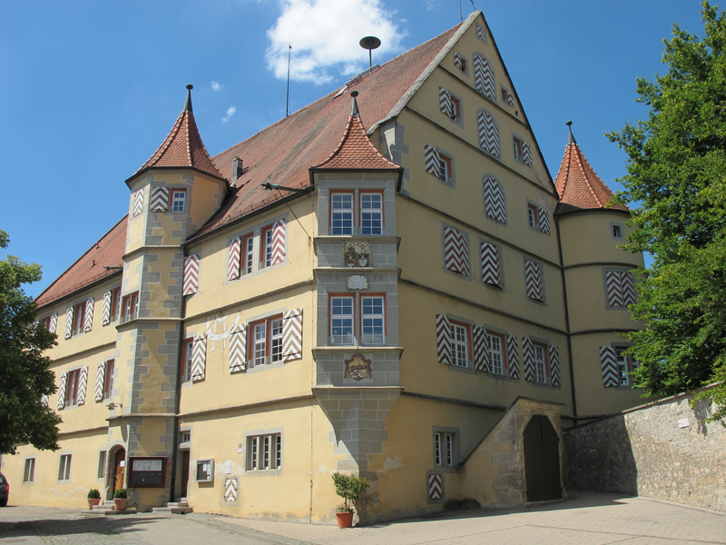 Ein historisches Gebäude mit zwei Türmen, rotem Ziegeldach und verzierten Fensterläden steht an einem sonnigen Tag vor einem blauen Himmel.
