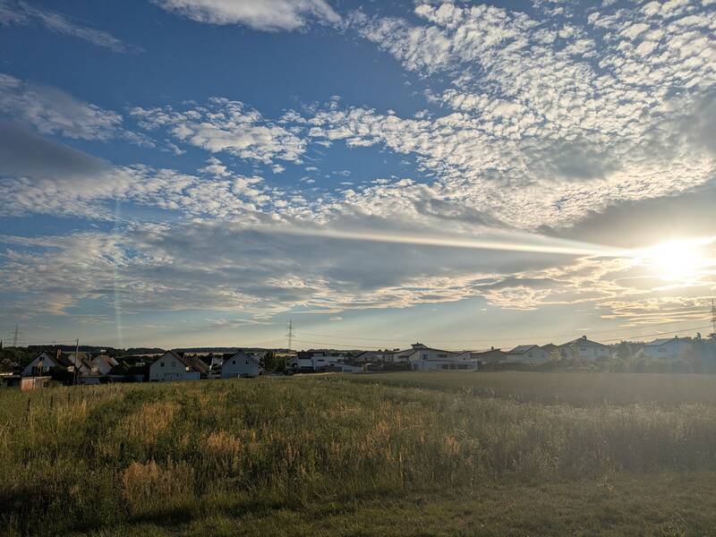 Eine weitläufige Wiese mit vereinzelten Häuserdächern im Hintergrund, unter einem Himmel mit dünner Wolkendecke und strahlendem Sonnenschein.