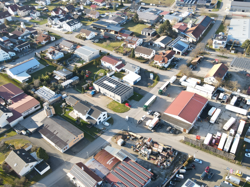 Eine Luftaufnahme zeigt ein Wohngebiet mit vielen Häusern und einigen Firmengebäuden, umgeben von geparkten Fahrzeugen und grünen Flächen.