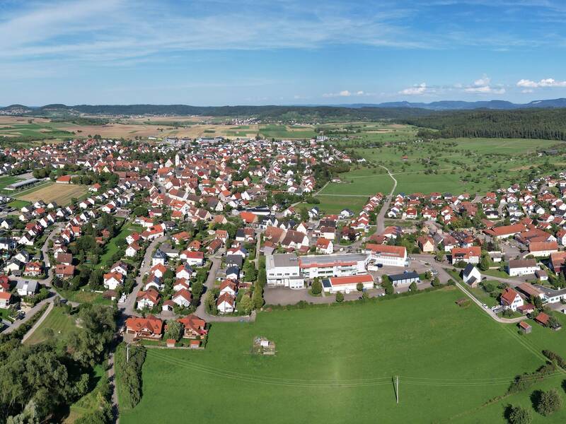 Eine weitläufige Luftaufnahme zeigt eine malerische, von Feldern und Wäldern umgebene Siedlung mit vielen Häusern und einer Kirche.