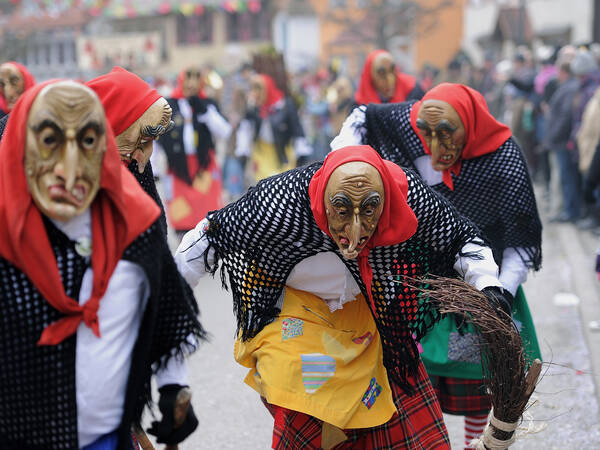 Auf dem Bild sind mehrere Personen in bunten, traditionellen Kostümen mit gruseligen Masken und roten Kopftüchern bei einem Straßenumzug zu sehen.
