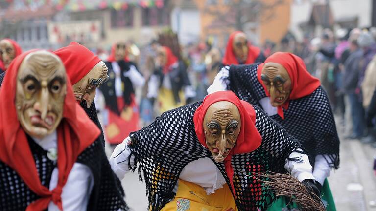 Auf dem Bild sind mehrere Personen in bunten, traditionellen Kostümen mit gruseligen Masken und roten Kopftüchern bei einem Straßenumzug zu sehen.