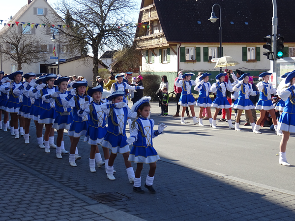 Eine Gruppe von jungen Menschen in blauen und weißen Kostümen zieht in einer Parade durch eine Straße, flankiert von Zuschauern und geschmückten Gebäuden im Hintergrund.