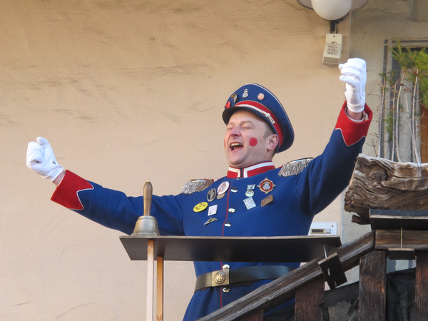 Ein Mann in einer blauen Uniform mit roten Verzierungen steht auf einer Bühne und hebt triumphierend die Arme, während er lächelt.