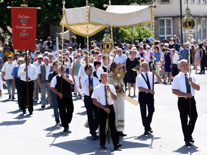 Eine Prozession zieht durch eine Straße, angeführt von Männern in weißen Hemden mit Krawatten, die ein Baldachin und religiöse Symbole tragen, während viele Menschen ihnen folgen.