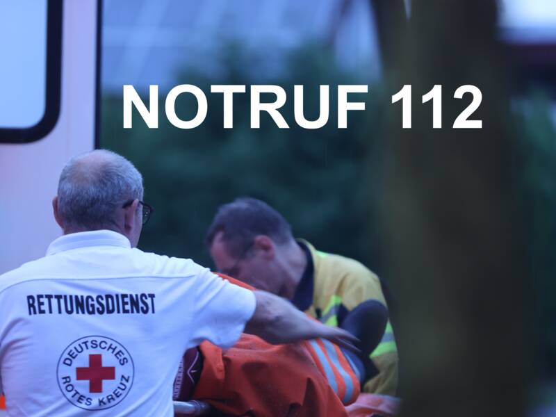 Ein Rettungsdienstmitarbeiter hilft einer Person auf einer Trage vor einem Krankenwagen, mit der Notrufnummer 112 im Bild.