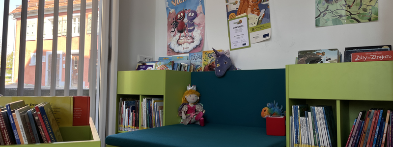 Ein gemütlicher Leseecke in einer Bibliothek mit grünen Regalen voller Kinderbücher, einer blauen Sitzbank und bunten Postern an der Wand.