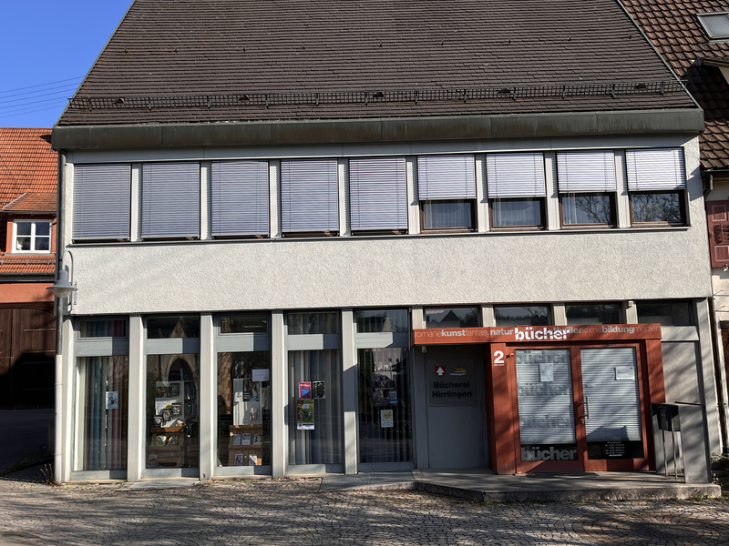 Das Bild zeigt die Front einer Bücherei mit großen Fenstern im Erdgeschoss und einem Schild mit der Aufschrift "Bücherei Hirrlngen".