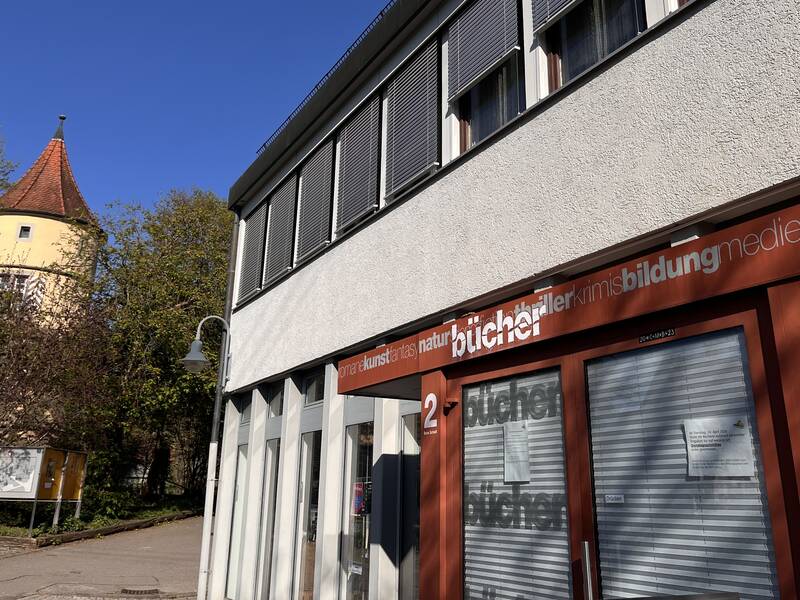 Ein modernes Gebäude mit der Aufschrift "bücher" auf einem roten Vordach steht neben einem runden Turm mit spitzem Dach und umgeben von Bäumen.