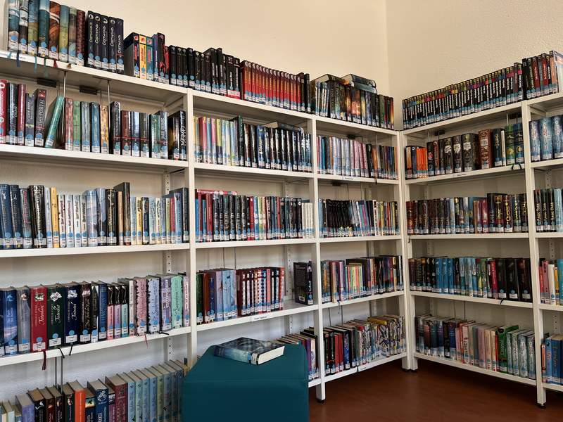 Ein Raum mit weißen Bücherregalen voller bunter Bücher in einer Bibliothek, mit einem Buch, das auf einem grünen Hocker liegt.