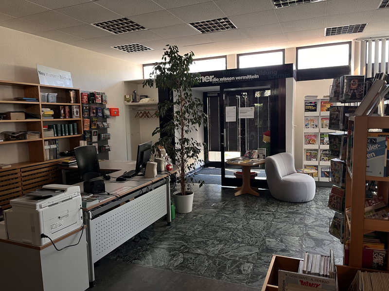 Ein heller Raum mit einem Schreibtisch, Regalen voller Bücher und Zeitschriften, einer Pflanze und einem gemütlichen Stuhl neben einer Glastür.