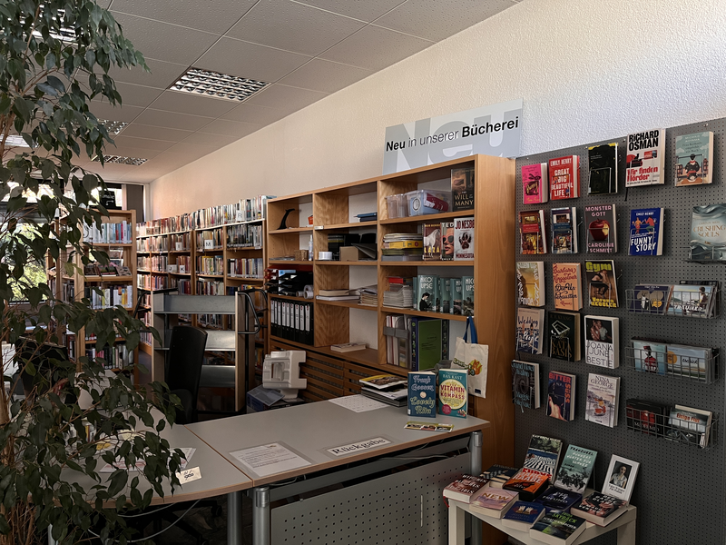 Ein Innenraum einer Bücherei mit vielen Bücherregalen, einem Schreibtisch und einem Regal für Neuerscheinungen.