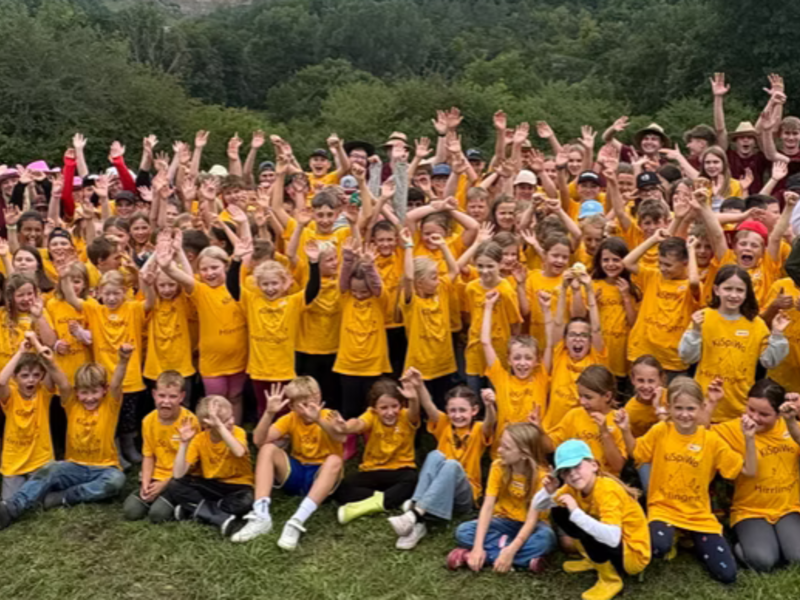 Eine große Gruppe von Kindern und Erwachsenen, die überwiegend gelbe T-Shirts tragen, jubelt draußen auf einer Wiese vor einer bewaldeten Landschaft.