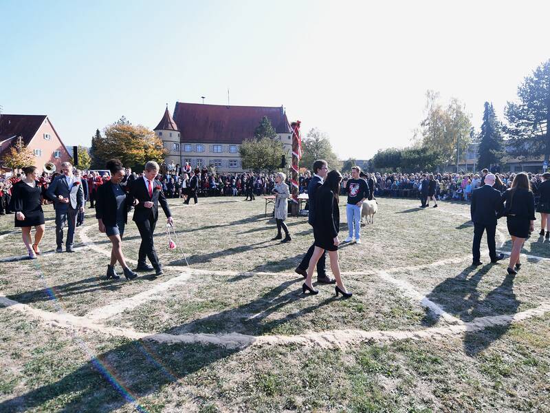 Eine Gruppe von Menschen in formeller Kleidung geht auf einer Rasenfläche, während im Hintergrund eine große Menschenmenge zuschaut und traditionelle Gebäude zu sehen sind.