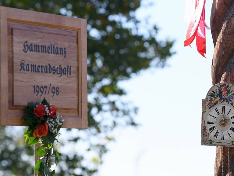 Ein Schild mit der Aufschrift "Hammeltanz Kameradschaft 1997/98" ist mit roten Rosen und grünem Laub dekoriert, daneben hängt eine verzierte Uhr mit floralen Mustern.