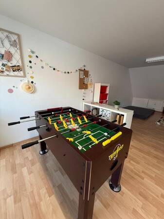 In einem Raum mit hellem Holzboden steht ein Kicker-Tisch neben einer weiß dekorierten Wand mit bunten Punkten und Pinnwand.