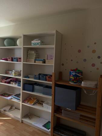 Ein heller Raum mit zwei weißen Regalen, die verschiedene bunte Boxen, Bücher und Bastelmaterialien enthalten, und daneben ein Holzregal mit einer großen grauen Box und einer Kiste mit Spielzeug.