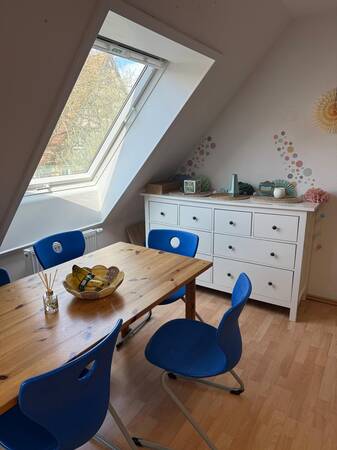 Ein helles Zimmer mit einem Holztisch, umgeben von blauen Stühlen, einem schrägen Dachfenster und einer weißen Kommode, dekoriert mit bunten Wandakzenten.