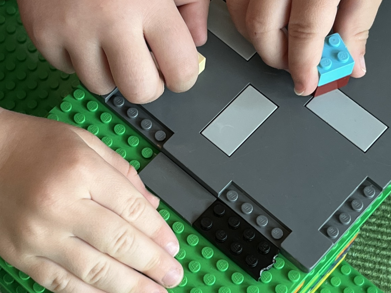 Kinder bauen auf einem grünen Teppich mit bunten Legosteinen eine Struktur, während eine Spielzeugfigur daneben steht.
