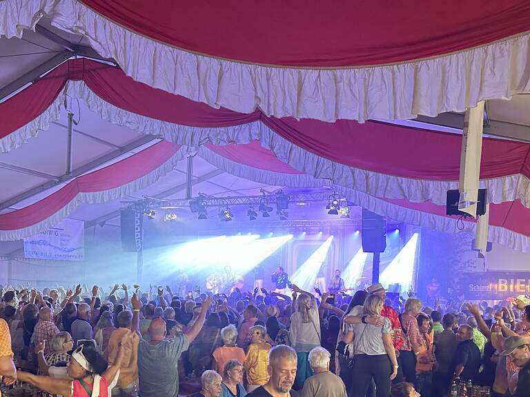 In einem großen Festzelt mit rot-weißer Dekoration feiern zahlreiche Menschen zu einer Bühne hin, auf der eine Musikband unter bunten Lichtern spielt.