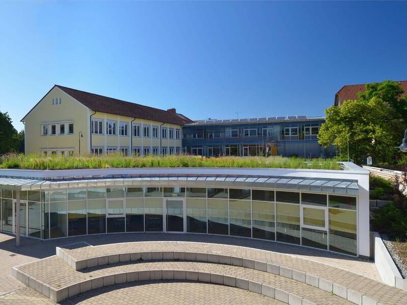 Das Bild zeigt ein modernes Gebäude mit großen Glasfenstern, einer terrassierten Grünfläche auf dem Dach und einem angrenzenden, gelben, mehrstöckigen Gebäude, umgeben von üppigem Grün und blauem Himmel.