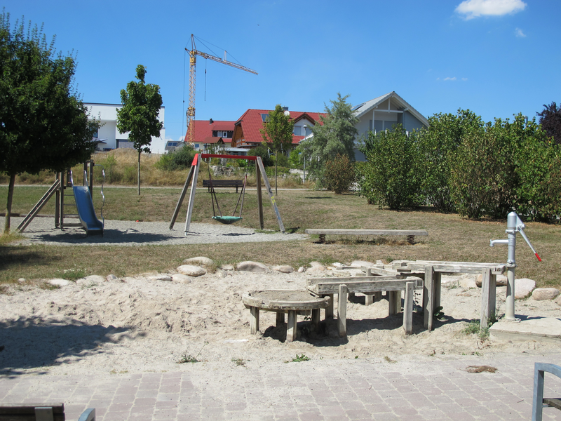 Ein Spielplatz mit einer metallenen Rutsche, einer Schaukel und einer Wasserpumpe befindet sich in einer grünen Umgebung mit Bäumen und einem Kran im Hintergrund.