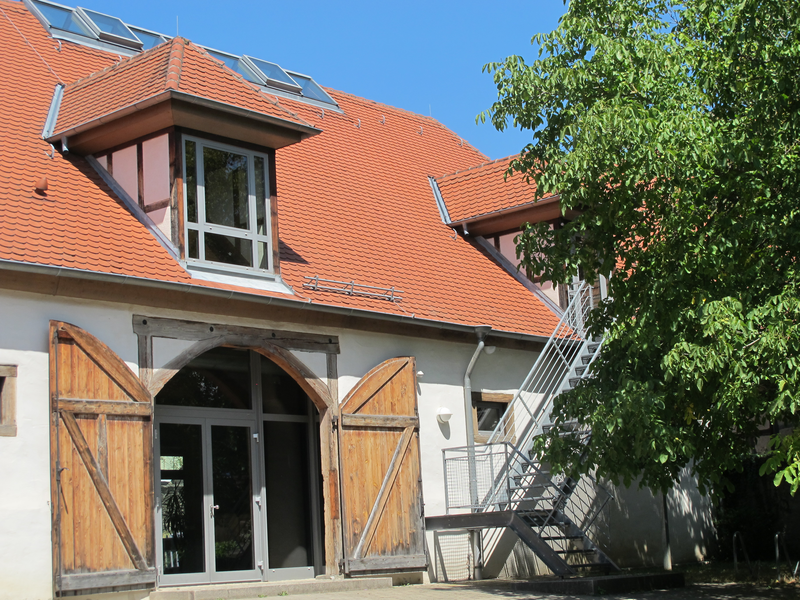 Ein Gebäude mit roten Dachziegeln, großen Holztüren, einem Dachfenster und einer Außentreppe, umgeben von einem Baum bei blauem Himmel.