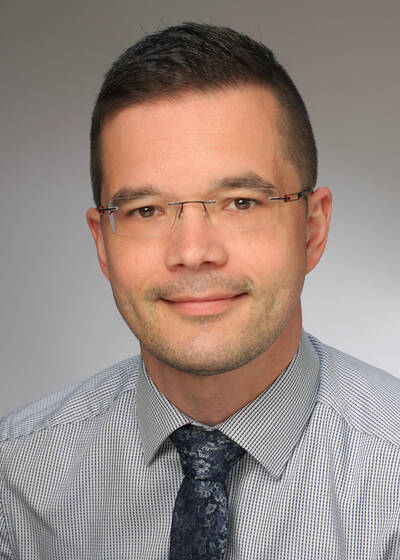 Ein Mann mit Brille und kurz geschnittenem Haar trägt ein kariertes Hemd und eine gemusterte Krawatte und lächelt leicht.