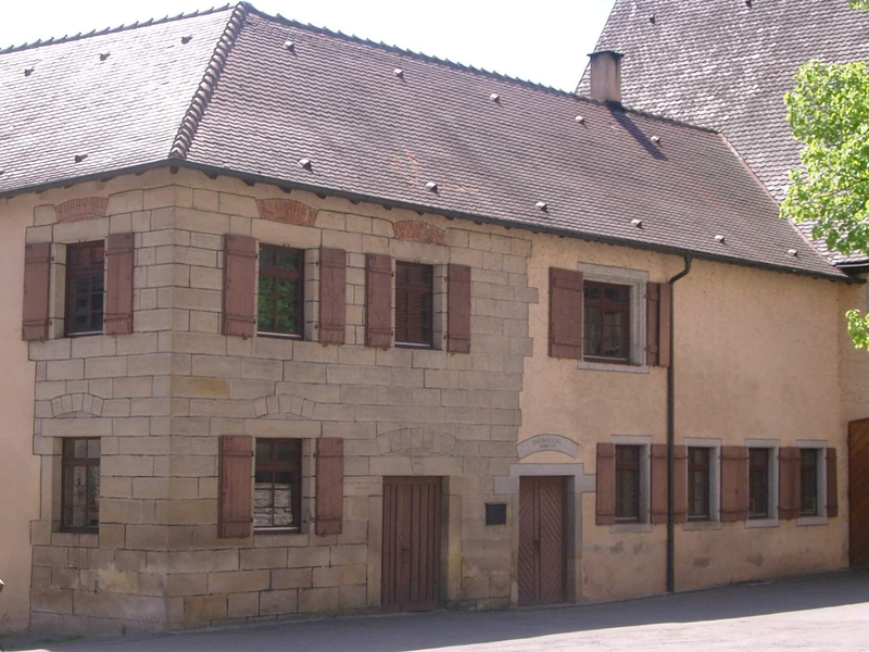 Ein zweistöckiges Gebäude mit hellen Steinwänden und dunkelbraunen Fensterläden, das unter einem großen Satteldach steht.