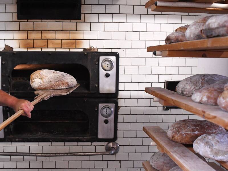 Ein Bäcker schiebt mit einer Holzschaufel einen Laib Brot in einen Ofen, während weitere Brote auf hölzernen Regalen daneben lagern.
