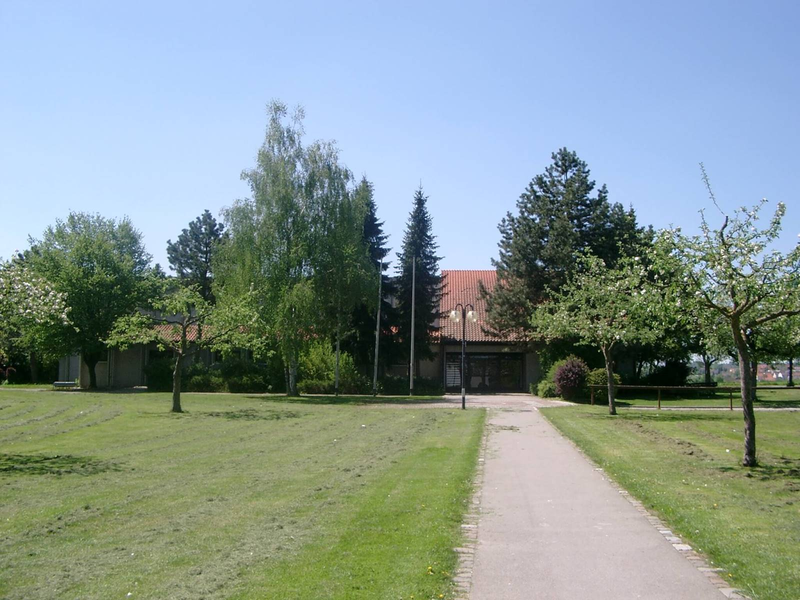 Ein gepflasterter Weg führt durch eine grüne Wiese zu einem Gebäude mit rotem Ziegeldach, das von hohen Bäumen umgeben ist.