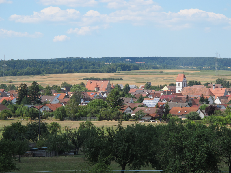 Blick auf ein malerisches Dorf mit roten Dächern und einer Kirche, umgeben von Wiesen und Bäumen unter einem leicht bewölkten Himmel.