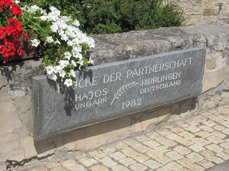 Ein Blumenbeet mit bunten Blumen befindet sich neben einer Mauer mit einer Gedenktafel zur Partnerschaft zwischen Hajós, Ungarn und Hirrlingen, Deutschland, datiert auf 1982.