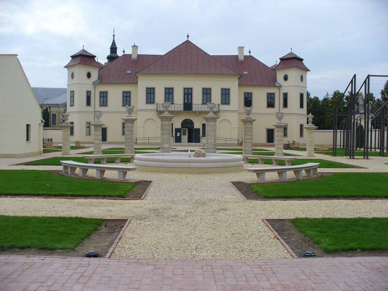 Ein elegantes, beige Schloss mit zwei Türmen steht hinter einem symmetrisch angelegten Garten mit einem runden Brunnen in der Mitte.