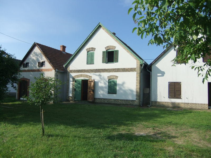 Drei weiße Landhäuser mit grünen Fensterläden und Schindeldächern stehen auf einem grasbewachsenen Grundstück unter einem klaren blauen Himmel.