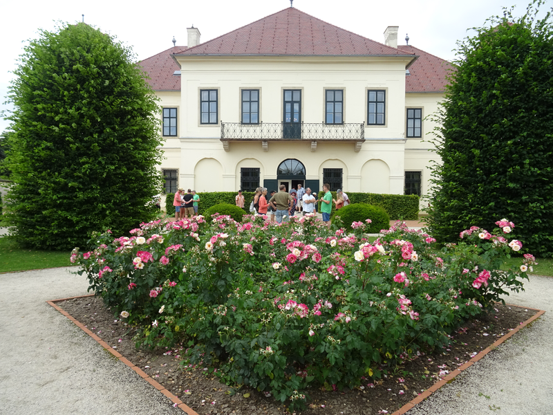 Vor einem großen, hellen Gebäude mit rotem Dach stehen mehrere Menschen im Freien, umgeben von einem gepflegten Garten mit blühenden rosa Rosen.