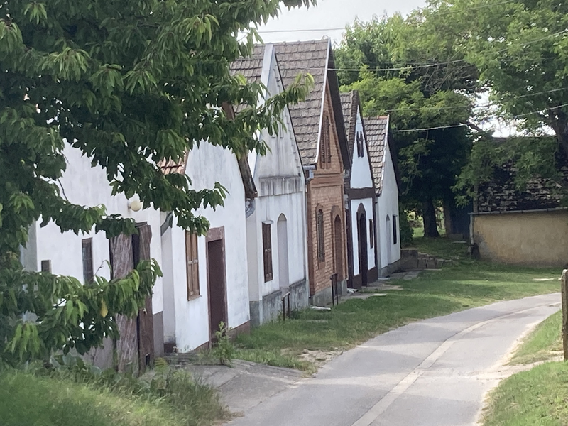 Eine Reihe von kleinen, traditionellen Häusern mit weißen und ziegelroten Fassaden steht entlang einer kurvigen, schmalen Straße, umgeben von grünen Bäumen.