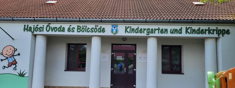 Das Bild zeigt die Fassade eines Gebäudes mit der Aufschrift "Hajósi Óvoda és Bölcsöde Kindergarten und Kinderkrippe," verziert mit einem Cartoon-Kind an der Wand.