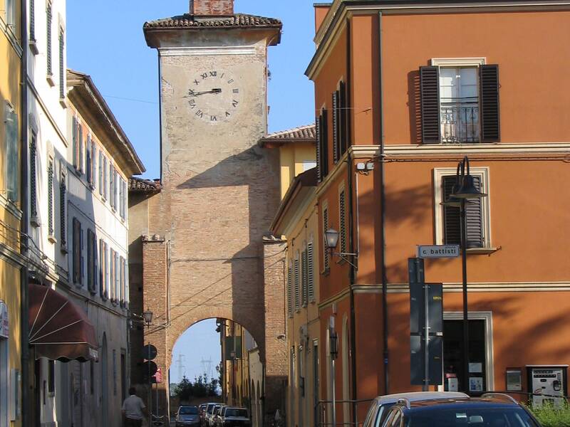 Eine belebte Straße in einer italienischen Stadt wird von einem historischen Turm mit Uhr und Glocke überragt, der zwischen zwei Gebäuden steht.