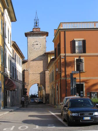 Eine belebte Straße in einer italienischen Stadt wird von einem historischen Turm mit Uhr und Glocke überragt, der zwischen zwei Gebäuden steht.
