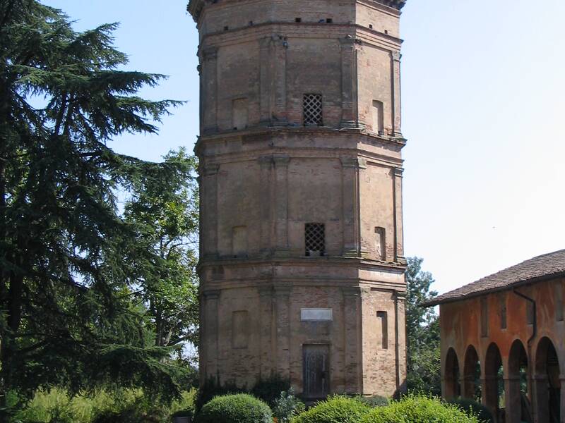 Ein hoher, dreistöckiger Ziegelturm mit einer Kuppel steht in einer grünen, gepflegten Landschaft neben einem Gebäudeflügel mit Arkaden.
