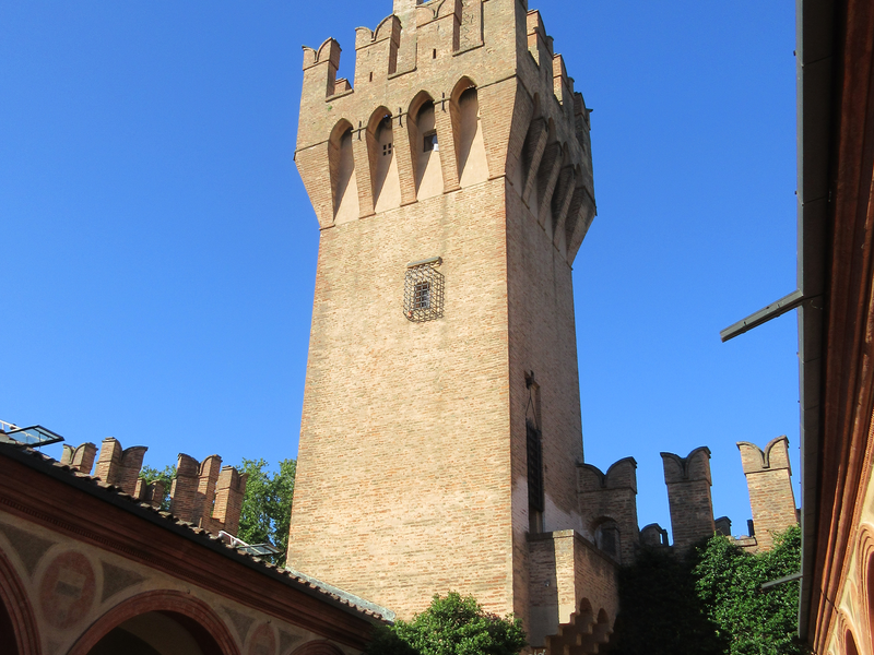 Ein hoher, mittelalterlicher Backsteinturm mit Zinnen erhebt sich in einem Innenhof vor einem klaren blauen Himmel.