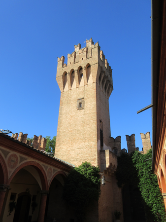 Ein hoher, mittelalterlicher Backsteinturm mit Zinnen erhebt sich in einem Innenhof vor einem klaren blauen Himmel.